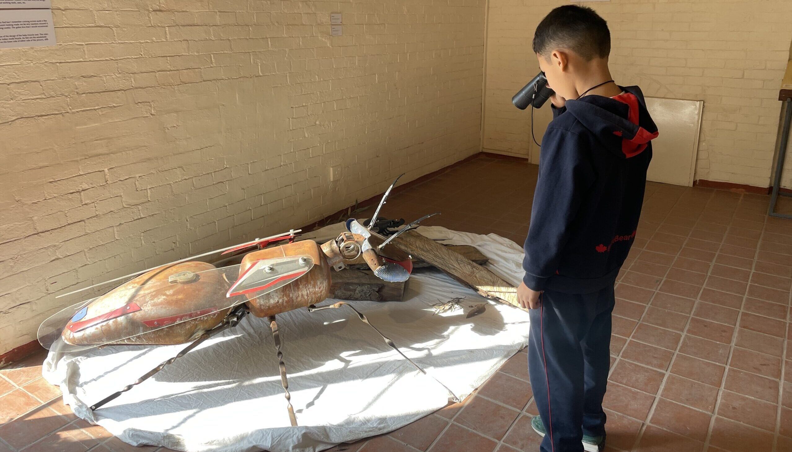 A young boy looks through his binoculars at a queen ant sculpture made out of waste materials like old motorcycle parts